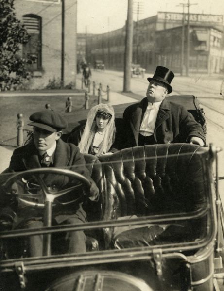 The Wall Street broker Mr. Berry (played by W.H. Brown) and his daughter (Billie West, wearing a hat with a white veil) sit in the back of a chauffeured open touring car in a scene still from "Above Par" (Reliance 1915).