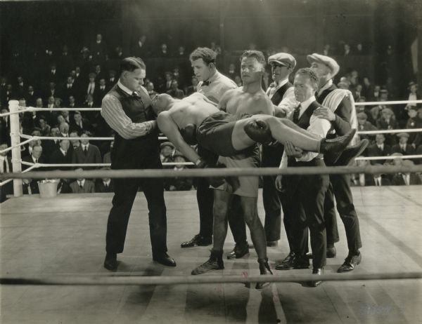 The boxer Pat Glendon, Jr. (played by Reginald Denny) carries his unconscious boxing opponent in his arms as the referee and various trainers gather around in the center of the ring in this scene still from "The Abysmal Brute" (Universal 1923).