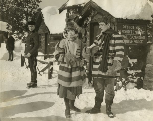 Royal Canadian Mounted Police Sergeant Flaherty (played by Tom Moore) looks on suspiciously as Jen Galbraith (Betty Compson) talks to Pretty Pierre (Jean De Briac) on a snowy street lined with log cabins, one of which is a RCMP office. Compson and De Briac both wear Hudson Bay blanket coats in this scene still from "Over the Border" (Lasky 1922).