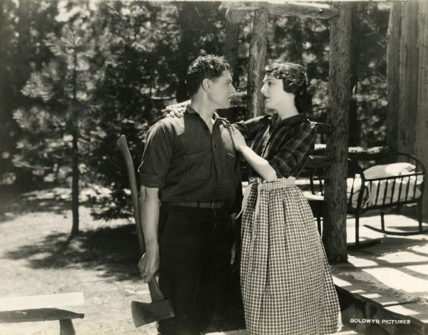 Forrest (played by John Bowers) holds an axe as Lilith (Leatrice Joy) gazes into his face in this scene still from the silent film "The Ace of Hearts." They are dressed in sturdy work clothes, plaids and woolens, and stand before a log cabin's porch on which is an empty baby cradle. Behind them are pine woods.
