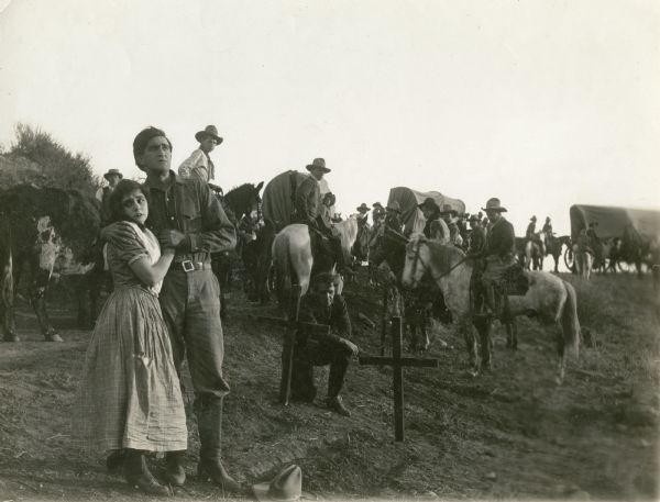 In the foreground Ethel Grandin and Francis Ford embrace at the foot of a grave. Behind them are three covered wagons, numerous men and women on horseback, and some cattle in this scene still from the one-reel silent film "Across the Plains," also known as "War on the Plains."
