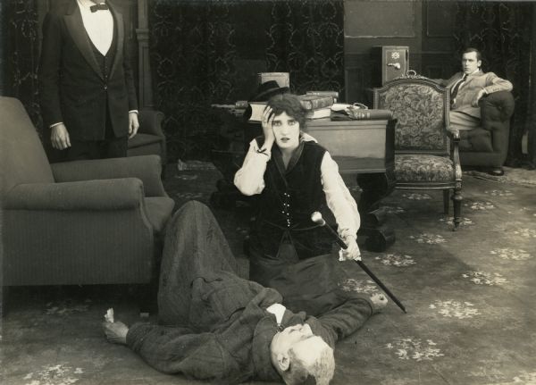 Mr. Pepper (played by Charles Lee) lies dead on the floor as his daughter Jane (Irene Hunt) kneels beside him in shock. In the background sitting by a small safe is her brother (Jack Conway) in this scene still from "Added Fuel," a two-part short silent film released May 15, 1915.