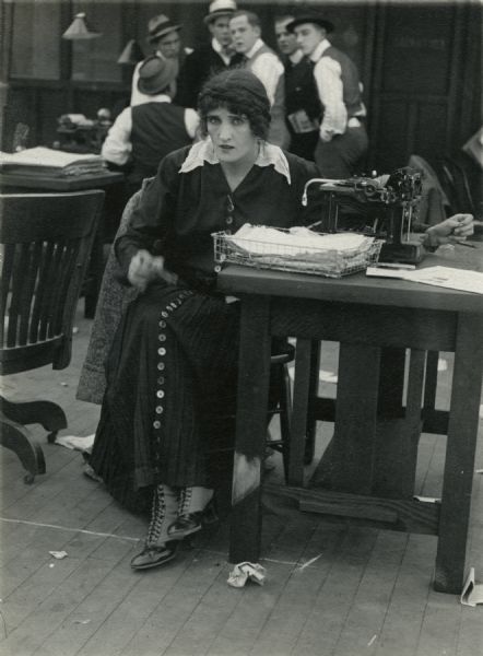 Jane Pepper (played by Irene Hunt) is a reporter for "The Daily Mail." She sits at her typewriter while a group of six men talk behind her back in this scene still from "Added Fuel" (Reliance 1915).