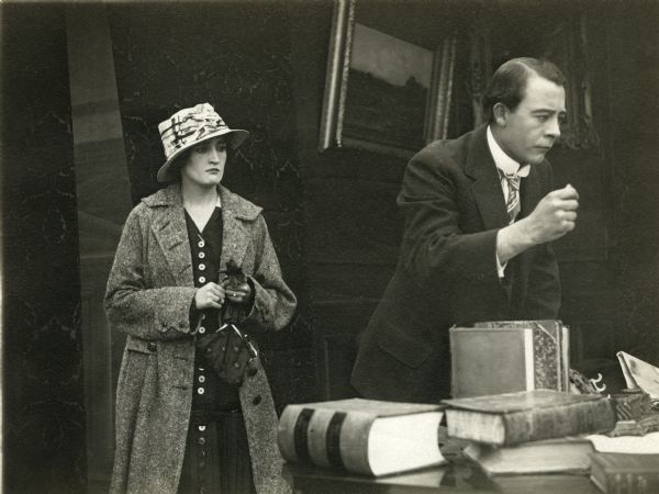 The actors Irene Hunt and Vester Pegg in a scene still from "Added Fuel." She wears street clothes, a heavy coat and hat, and carries a purse. He is behind a desk scattered with large legal books.