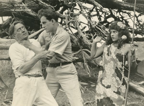 Templeton (played by Jack Perrin) throttles a man as Marama Thurston (Edith Roberts) looks on in alarm in this scene still for "The Adorable Savage" (Universal 1920). She wears a muumuu and shell necklace and holds a bamboo pole.