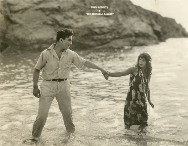 Templeton (played by Jack Perrin) holds Marama Thurston (Edith Roberts) by the wrist as they stand calf-deep in the surf on the island of Fiji in a scene still from "The Adorable Savage" (Universal 1920). He is dressed in khaki pants and shirt, she in a muumuu with a shell necklace.