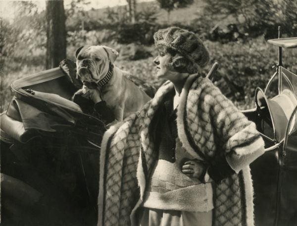 The actress Corinne Griffith posing with an English bulldog and an open one seat runabout automobile in a publicity still for the silent film "The Adventure Shop."