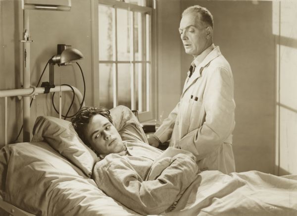Still from the 1950 United Artists film "The Men," featuring Marlon Brando (as Ken Wilocek) and Everett Sloane (as Dr. Brock). 

The typewritten caption reads: "Bedside Manner Gets Tough - Everett Sloane, in the role of Dr. Brock, gets tough with patient Marlon Brando in an effort to make him work toward recovery, in the Stanley Kramer production, "The Men," starring Brando and Teresa Wright under the direction of Fred Zinnemann."