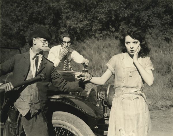 Still from the 1922 Hodkinson film "Affinities," featuring Pietro Sosso (as Professor Savage), John Bowers (as Day Illington), and Colleen Moore (as Fanny Illington). A man dressed as a sherriff (probably Pietro Sosso) restrains Colleen Moore by the wrist in front of the hood of an automobile. Beyond the car a young man (perhaps John Bowers) looks on.