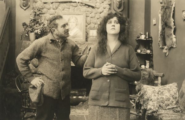 Still from the 1916 Vitagraph film "Tried for His Own Murder," featuring Maurice Costello (playing Ransford) and Leah Baird (playing Irene Gardner). They stand in a rustic interior with a gun over the fireplace and an animal skin on the wall.