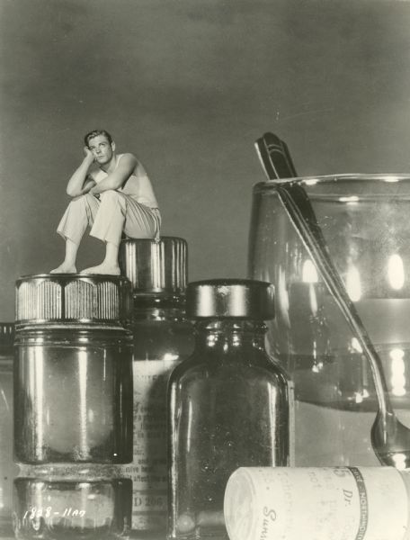 Still from the 1957 Universal film "The Incredible Shrinking Man," featuring Grant Williams (as Scott Carey), a tiny man sitting on top of medicine bottles on a bedside table. The caption on the back of the print reads: "Grant Williams, deminished [sic] to less than an inch in height by a mystery ailment, finds that the everyday world he knew has become a gargantuan chamber of horrors in which he is trapped in Universal-International's terrifying science-fiction thriller, 'The Incredible Shrinking Man,' which stars Williams with Randy Stuart."