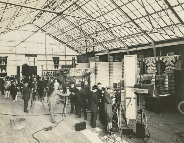 Production still showing how three or more silent film scenes could be simultaneously filmed under the glass roof of the Thanhouser studio. The print is captioned "Thanhouser 2nd Studio," suggesting that this is the interior of the Thanhouser studio built on Main Street in New Rochelle, New Jersey, to replace the first studio which burned in 1913. Visible are actors in costume, directors, cameramen, sets, a Klieglight, and four Pathé silent film cameras.