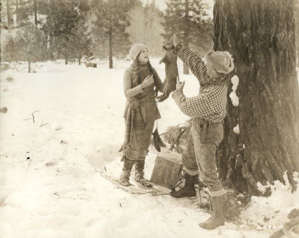 Snowy outdoor location scene still from the 1919 Famous Players silent film "A Daughter of the Wolf," featuring Lila Lee (playing Annette Ainsworth) and Clarence Geldart (playing "Wolf" Aisnworth). The trapper is showing his daughter a rabbit he has caught and she is overjoyed. Both are dressed in heavy winter clothes: he wears a fur hat, bulky checked shirt, and logging boots; she is covered in layers of woolen knitwear and wears snowshoes.