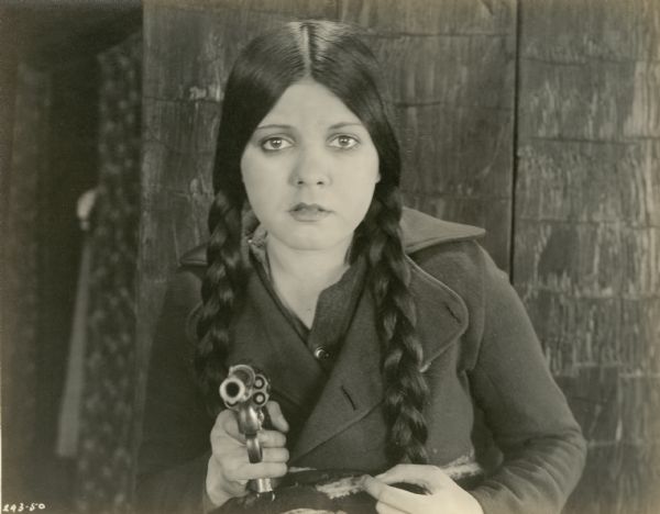 Lila Lee (as Annette Ainsworth) confronts the camera with a loaded revolver in a scene still from the 1919 Famous Players silent film "A Daughter of the Wolf." Her hair is in braided pigtails.