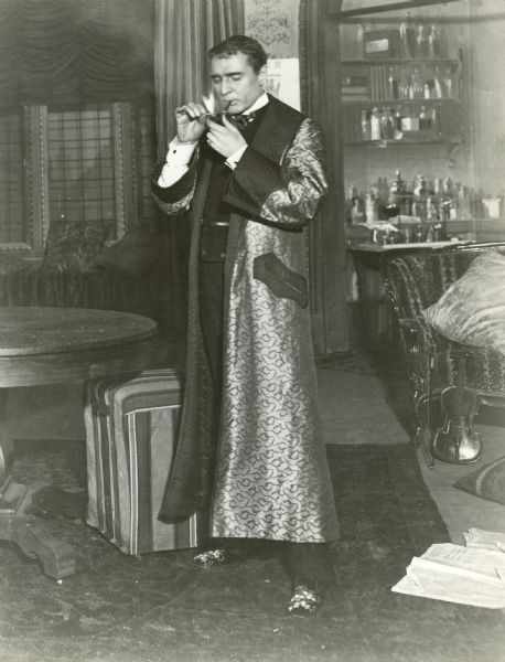 Publicity still of William Gillette in costume as Sherlock Holmes for the 1916 Essanay silent film production "Sherlock Holmes," based upon the play Gillette himself wrote in 1899. He stands in a dressing gown and lights his pipe with a match. Behind him are a violin leaning against a sofa and chemical apparatus in an alcove.