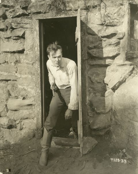 Royal Beaudry (played by Charles Ray) peers cautious from the open door of a rough stone building with a revolver in his hand in a scene still from "The Sheriff's Son" (Paramount, 1919).