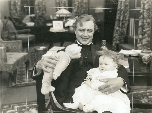 The Reverend Eric Norton (played by Hobart Bosworth) entertains a baby with a teddy bear in a publicity still from "The Scarlet Sin" (Universal, 1915).