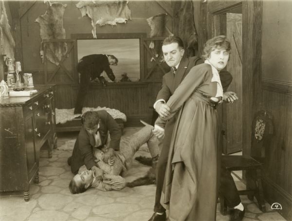 Kitty Gray (played by Grace Cunard) holds a knife and struggles with a ruffian, while behind them two men fight on the floor and a third escapes out the window in a scene still from episode 5 "The Underground Foe" of the Universal serial "The Broken Coin." They are in a rustic building in the mountains with animal pelts on the walls and beer steins on a sideboard.