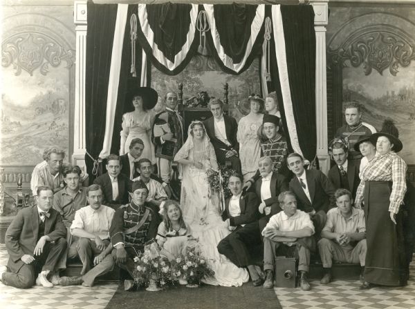 Group shot of the cast and crew for the Universal serial "The Broken Coin," taken after shooting the final scene. Grace Cunard and Francis Ford are in the center on the throne, the young actress Gertrude Short is seated on the floor in front of Ms. Cunard, and John Ford is third from the left. A Pathé motion picture camera is on the right in front of a man who most probably is Harry McGuire Stanley, the cameraman.
