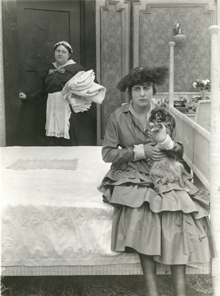 Grace Cunard sits on a bed and frets while a maid looks on in a publicity still for the Universal serial "The Broken Coin" for which Cunard was both writer and leading lady. Cunard clutches a Pekingese dog.<p>The original caption on the back of the print reads: "Grace Cunard in her house and a quandary over the subject matter which she has to write into an additional series installment of 'The Broken Coin.'"