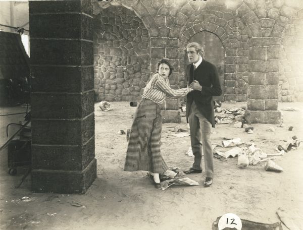 Kitty Gray (played by Grace Cunard) and Count Frederick (played by Francis Ford) stand in a dungeon and stare off-camera in horror in a scene still from Episode 12 "A Cry in the Dark" of the 1915 Universal serial "The Broken Coin." Amid some rubbish on the floor behind them is a skeleton.