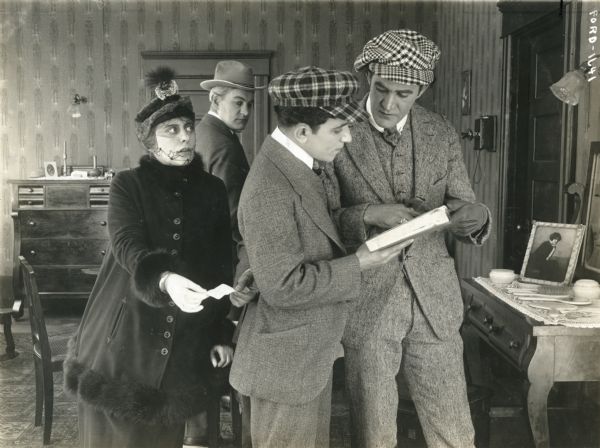 Grace Cunard (playing Kitty Grey) slips a note to Harry Mann as he examines a book with Francis Ford (playing Count Frederick) in a scene still from the Universal serial "The Broken Coin." They are in a bedroom with a telephone on the wall and dressing table laid out with combs, brushes, and make-up.