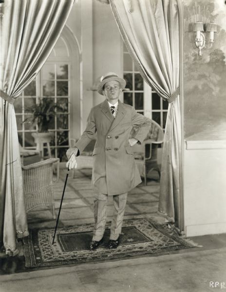 Speck Brown (played by Wesley Barry) wearing fancy new city clothes--a Chesterfield coat, checked trousers, spats, cane, white gloves, and a straw boater--stands in a doorway in a scene still for "School Days" (Warner, 1921).