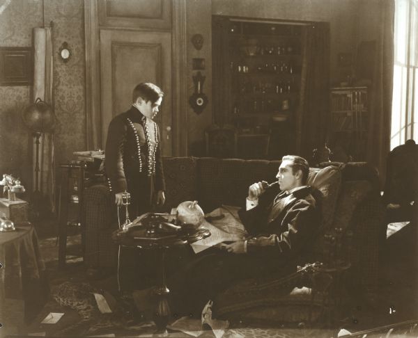 Billy the pageboy (played by Jerry Devine) talks to Sherlock Holmes (John Barrymore), who is stretched out on a sofa smoking a pipe in a scene still from the silent "Sherlock Holmes" (Goldwyn 1922).