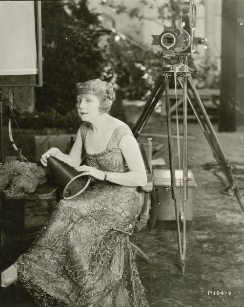 Actress Kathlyn Williams wearing an evening dress sits in William C. de Mille's director's chair holding his small megaphone. Behind her is one of the Bell & Howell model 2709 silent film cameras used to photograph "Conrad in Quest of His Youth," a Famous Players-Lasky production of 1920.