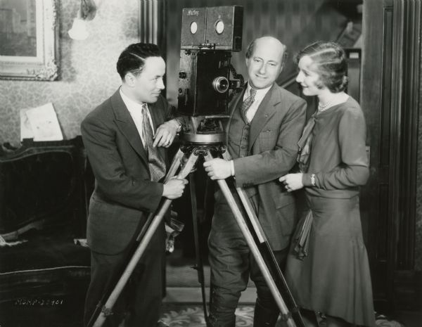 J. Peverell Marley, cameraman (left), and Cecil B. DeMille, director (right), pose beside a Pathé motion picture camera mounted on a large tripod. An unidentified woman stands with them.