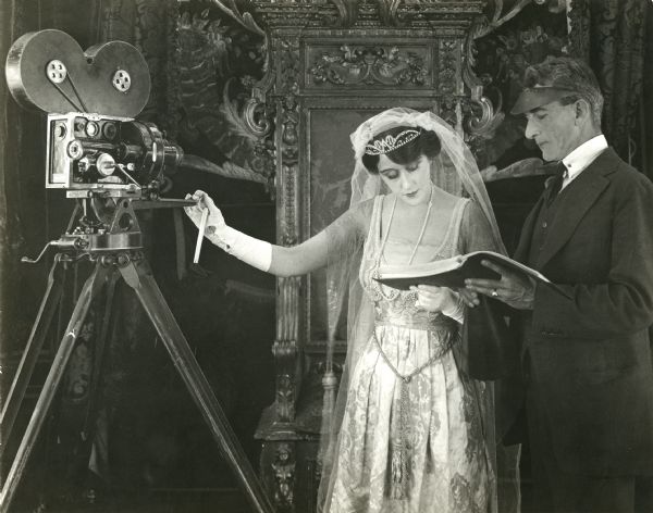 Production still of Olga Petrova and her director George Irving looking over the script for "Daughter of Destiny." Next to them is a Bell & Howell model 2709 motion picture camera on its tripod. Petrova is in a regal satin gown with veil, tiara, and long white gloves. Irving wears a suit and a visor.
