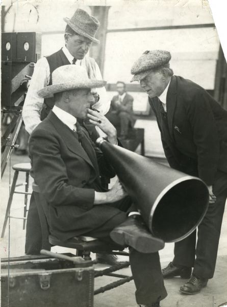 Director D.W. Griffith, seated with straw hat and large megaphone, confers with scriptwriter Frank E. Woods (bending, wearing a cloth cap), and cameraman G.W. Bitzer in a production still from "Intolerance." Behind Bitzer is a Pathé camera.