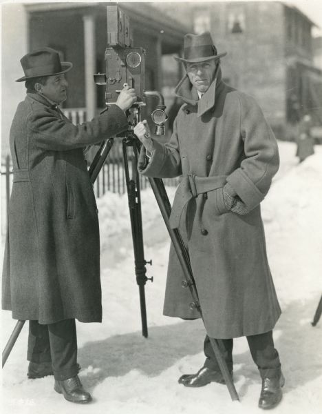 Cameraman G.W. Bitzer and director D.W. Griffith stand in the snow on a New Jersey street in a publicity still for "Way Down East." They are posed with Bitzer's Pathé camera which has a bicycle lamp attached to it by a tube. This was Bitzer's method of avoiding static discharges inside the camera, a problem in cold, dry weather.