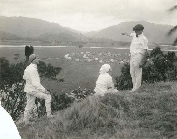 From left to right: Cameraman J. Roy Hunt with a Pathé camera, Annette Kellerman (seated), and director Herbert Brenon pointing to a caravan scene in the valley below for the 1916 Fox Film Corporation silent "The Daughter of the Gods" which was filmed in Jamaica. They are dressed in tropical whites.