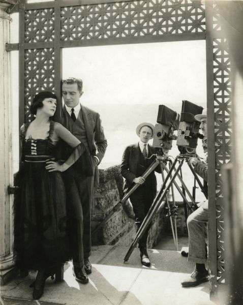 Original caption: "Photographing Norma Talmadge on the Highest Spot in New York"
"The patient still man has caught Norma Talmadge and her leading man Eugene O'Brien on location on the Billings estate at 194th St., and Wadsworth Ave. This estate is on the highest point in New York City overlooking the Hudson. Director Sidney A. Franklin unlike most directors is trying to hide from the still camera. The gentleman whose Mephistolean countenance is barely visible is Alfred Moses and his partner in the centre is Edward Wynard, both cameramen for Miss Talmadge."