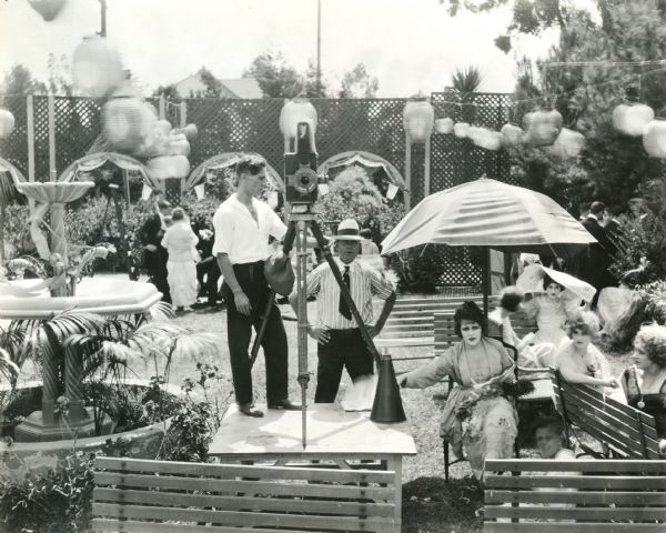 Ruth Roland sits under an unbrella in a garden scene with actors and motion picture crew members around her. A Pathé camera stands on a platform.