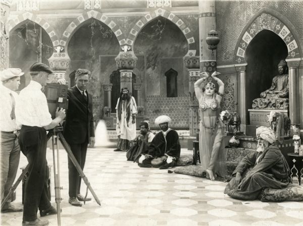 Mack Sennett, in a dark suit, and two technicians stand by a Prestwich camera on the set of an unidentified Middle Eastern themed Keystone Comedy. A belly dancer strikes a provocative pose, and four men are costumed as Arabs. Strangely enough, there is also a Buddha in an alcove.