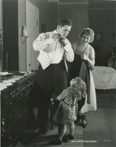 Tom Donaldson (played by John Bowers) is dressing with the help of his wife Grace (Louise Lovely) in a scene still from "The Poverty of Riches" (Goldwyn 1921). The little girl looking up at him is probably Barbara Maier, who played his daughter in the film.