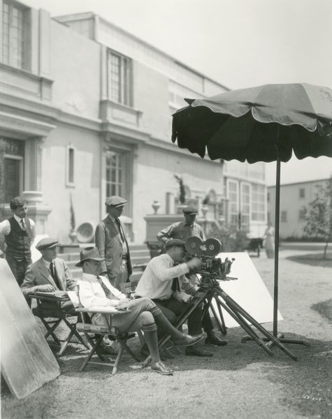 The director (Reginald Barker, seated wearing knickers and over-the-calf socks), cameraman cranking a Bell & Howell model 2709 (most probably Percy Hilburn), and other crew members watch as a scene is being shot outdoors.