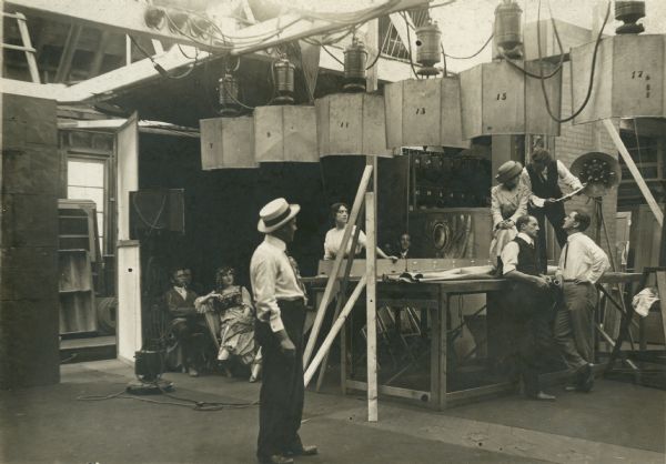 Florence La Badie (sitting on stage in light coat and hat), Carl Leviness (leaning against stage with megaphone), and Harry Benham (far right looking up at La Badie) in a production still from an unidentified Thanhouser silent film.