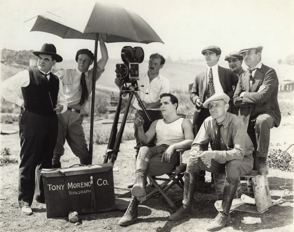 Antonio Moreno in make-up, jodhpurs, and an undershirt, sits under an umbrella along with a cameraman and a Bell & Howell camera with other cast and crew members in a production still. The camera case at his feet is labeled "Tony Moreno Co. Vitagraph."