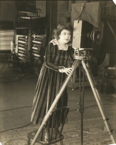 Marguerite Clark looks through the viewfinder of a Pathé motion picture camera in a publicity still.