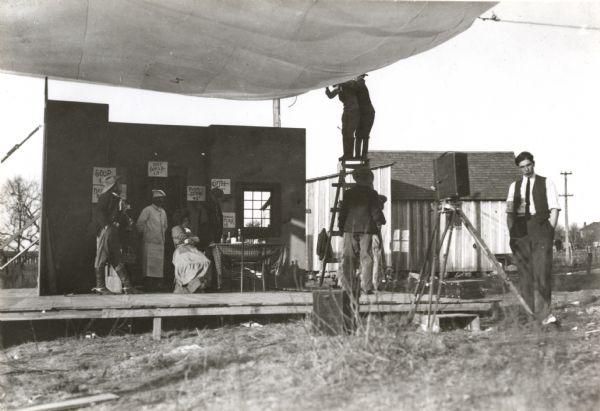 An outdoor studio used by the Eclair American Company while making silent films. Shown are four or five actors in a restaurant set on a low platform covered by a canvas diffuser. Facing them, on a tripod, is an English-style motion picture camera, perhaps a Williamson.