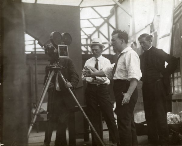 Thomas H. Ince gestures to a cameraman who is moving a Bell & Howell camera on its tripod. Over Ince's shoulder is William S. Hart in costume as a minister for an unidentified silent film production.