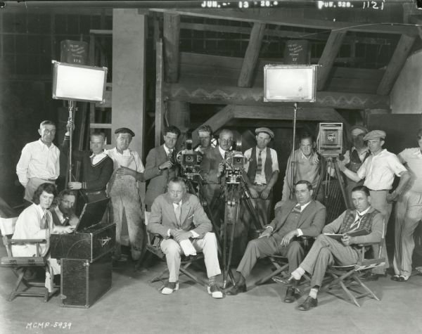 Director Nick Grinde (in two-tone shoes) and 15 members of his film crew pose for a MGM production still. Present are a woman playing the harmonium (far left), a still photographer and his 8 x 10 inch view camera (right), carpenters, painters, lighting technicians, and two motion picture cameramen. Directly behind Grinde is a Bell & Howell model 2709 camera, probably cranked by George Gordon Nogle, and to the right, a Mitchell camera probably cranked by Arthur Reed. The film may have been "Beyond the Sierras."