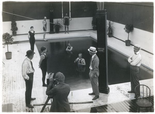 Production still of the Keystone pool. On the far left is Mack Sennett, coatless, in derby and suspenders. On the far right is Adam Kessel, coatless, in a straw hat. In the middle a cameraman cranks a Moy & Bastie motion picture camera. The nearest swimmer is Al St. John.