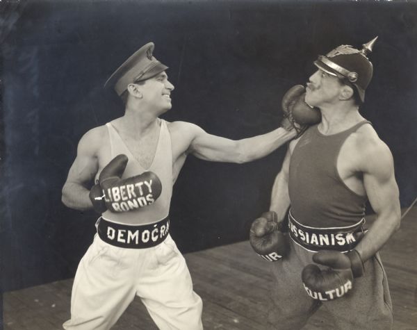 Democracy (played by Douglas Fairbanks) punches Prussianism (Bull Montana) on the jaw in a promotional still from "Sic 'Em, Sam" a short silent film made for the Fourth Liberty Loan Drive in World War I. Fairbanks wears a U.S. Army officer's visor cap, light-colored tank top and jodhpurs, and boxing gloves labeled "War Bonds." Montana, in a Kaiser Wilhelm moustache, wears a dark tank top and jodhpurs, a pickelhaube, and gloves labeled "Kultur."