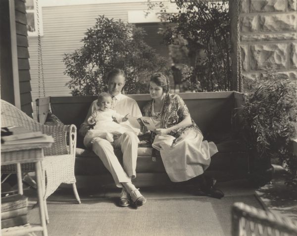 Silent film star Conrad Nagel sits on a porch swing with his infant daughter Ruth Margaret Nagel on his knee and his wife Ruth Helms by his side reading a book.
