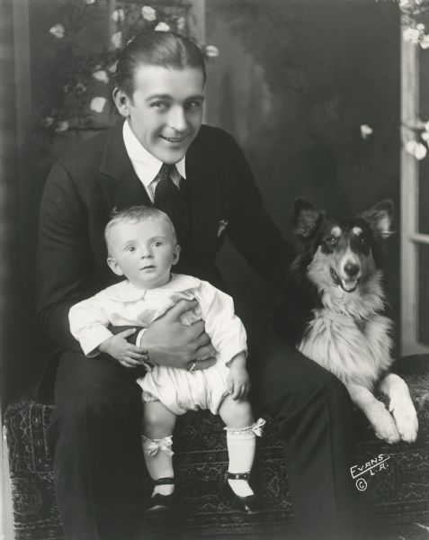 Silent film star Wallace Reid with his infant son Wallace Reid, Jr., on his knee and a collie dog.