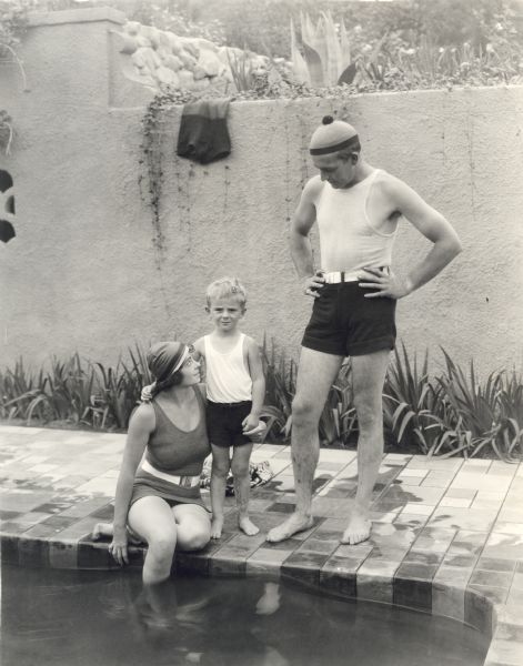 Dorothy Davenport Reid, Wallace Reid, Jr., and the silent film star Wallace Reid in bathing suits at the edge of a swimming pool.